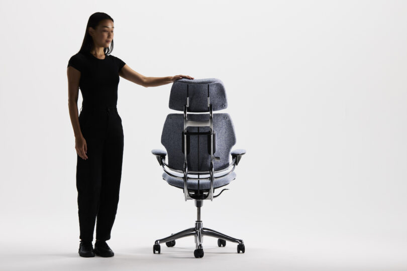 A person in black stands beside a modern ergonomic office chair with a unique backrest design on a white background