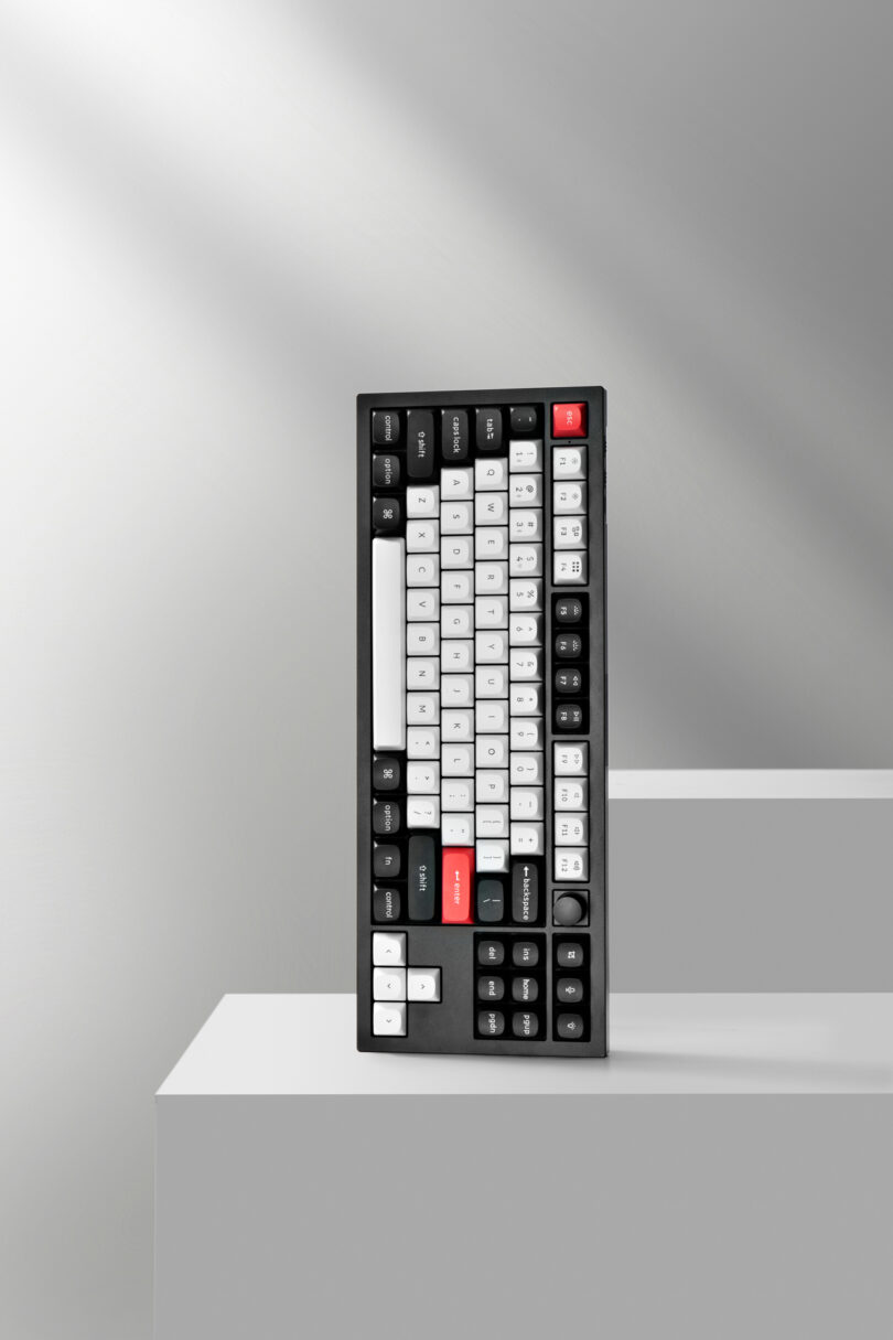 A black Keychron mechanical keyboard with white and black keys, a red escape key, and colorful accents is placed upright on a white geometric surface.