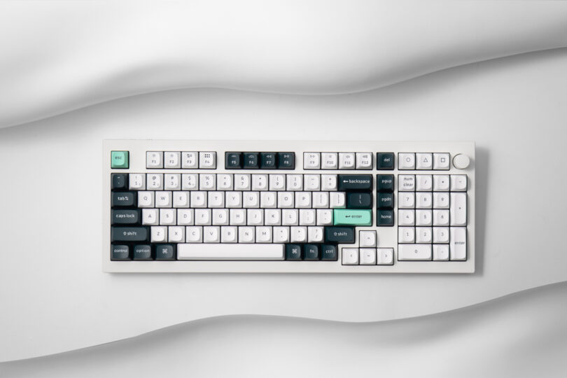 A Keychron mechanical keyboard with a white frame, featuring black, white, and green keycaps, is placed against a white, wavy textured background.