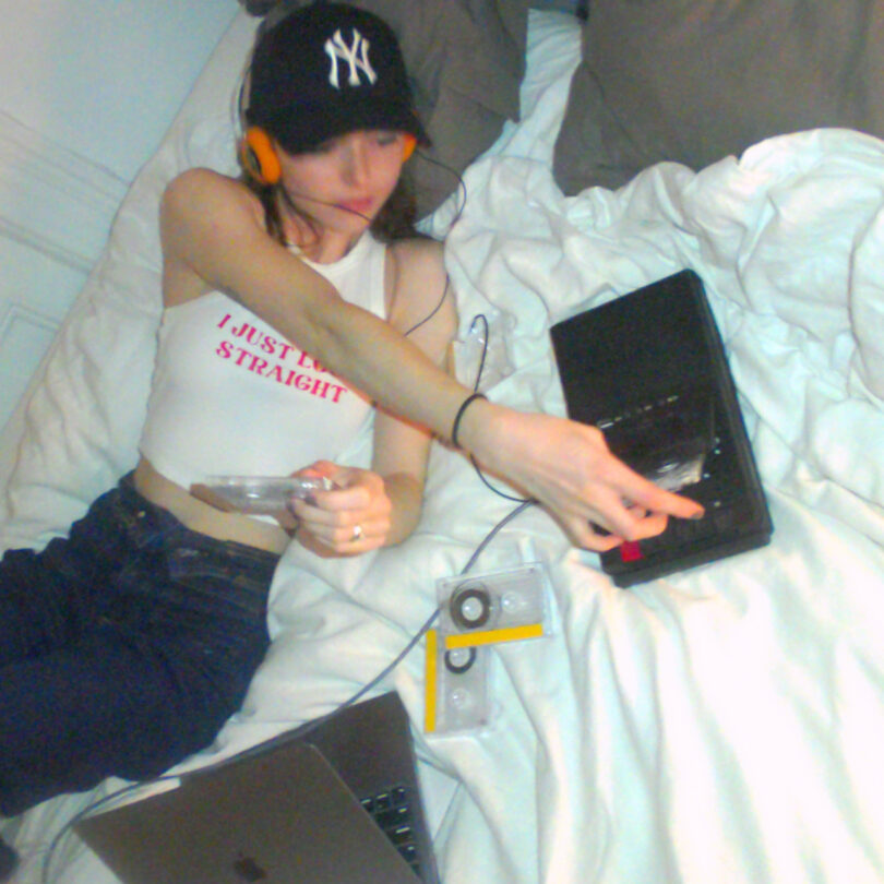 Person in a baseball cap lies on a bed, listening to a cassette player with headphones. Nearby are cassettes and a laptop.