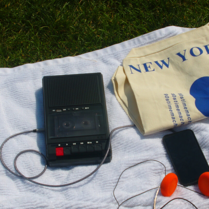 A portable cassette player, earphones, and a smartphone are placed on a white cloth next to a tote bag on grass.