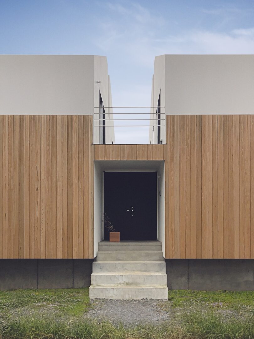 Modern minimalist building with wood paneling, white upper facade, and a black entrance door at the top of concrete steps.