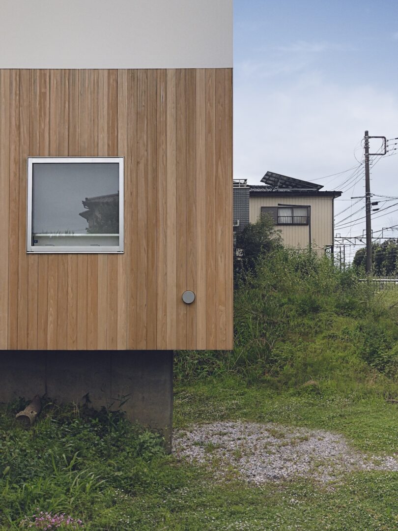 A wooden building with a single square window stands next to a grassy area, with another building and power lines in the background under a partly cloudy sky.