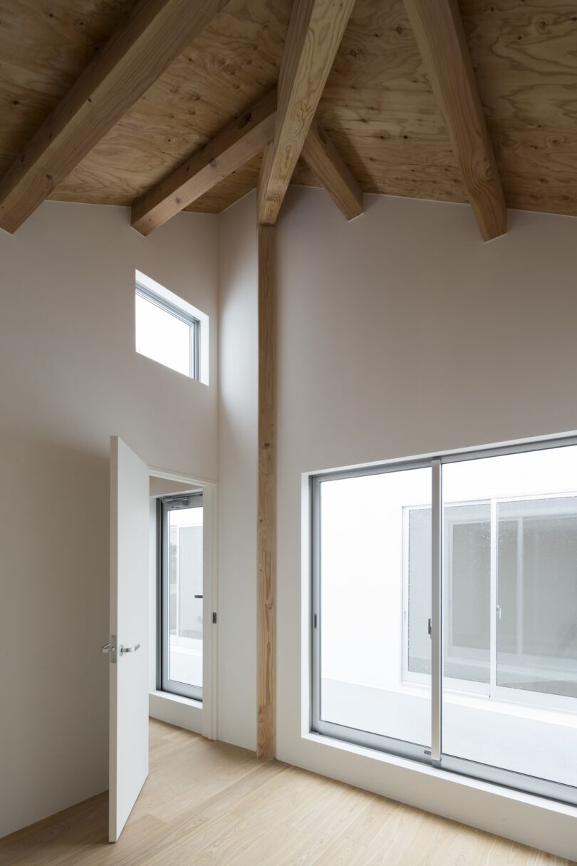 Minimalist interior with wood beam ceiling, open door, large windows, and light wooden floor, creating a spacious and bright atmosphere.