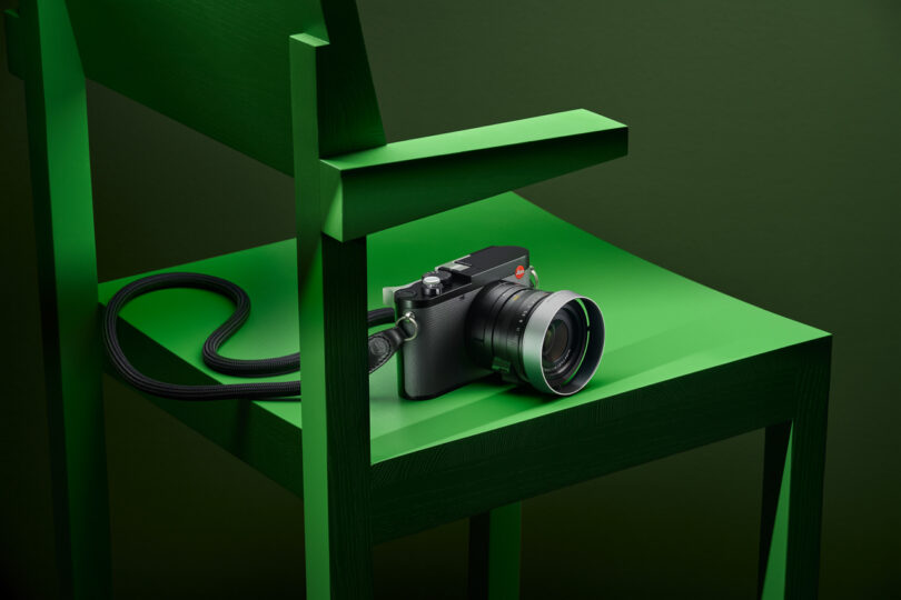 A black camera sits on the seat of a green wooden chair against a green background.