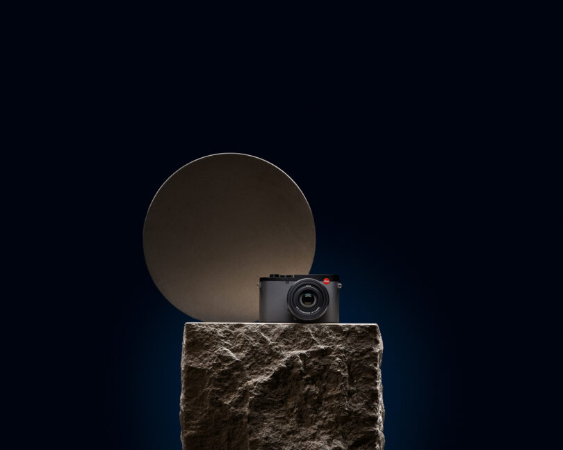 A camera sits on a stone pedestal with a circular object in the background against a dark backdrop.