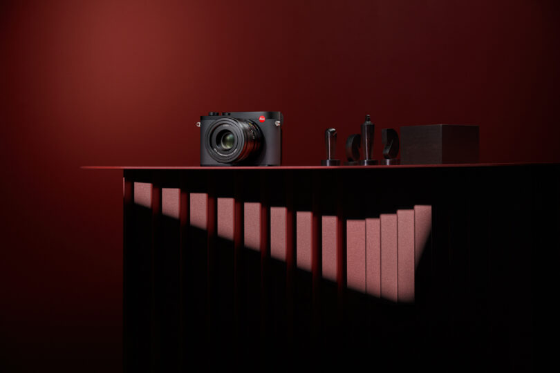 A black camera and accessories are displayed on a red table against a dark red background, illuminated by a diagonal beam of light.