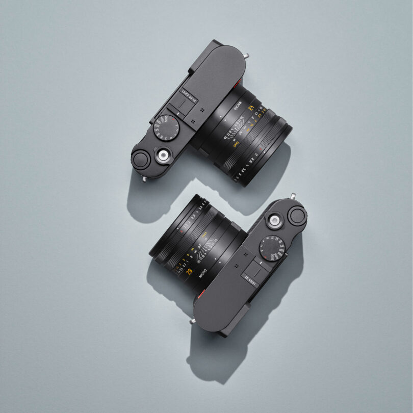 Two black cameras with attached lenses are positioned diagonally on a light gray surface.