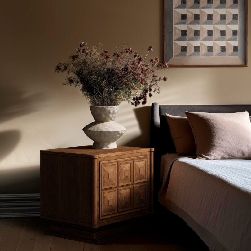 Bedroom scene with a wooden bedside table, a geometric vase of dried flowers, and a bed with pink pillows. A framed geometric artwork hangs on the wall