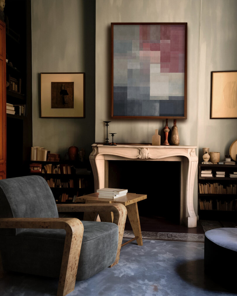 A cozy room with a gray armchair, a wooden table, and a fireplace. Abstract and framed art hang above the fireplace, surrounded by bookshelves filled with books and decorative item