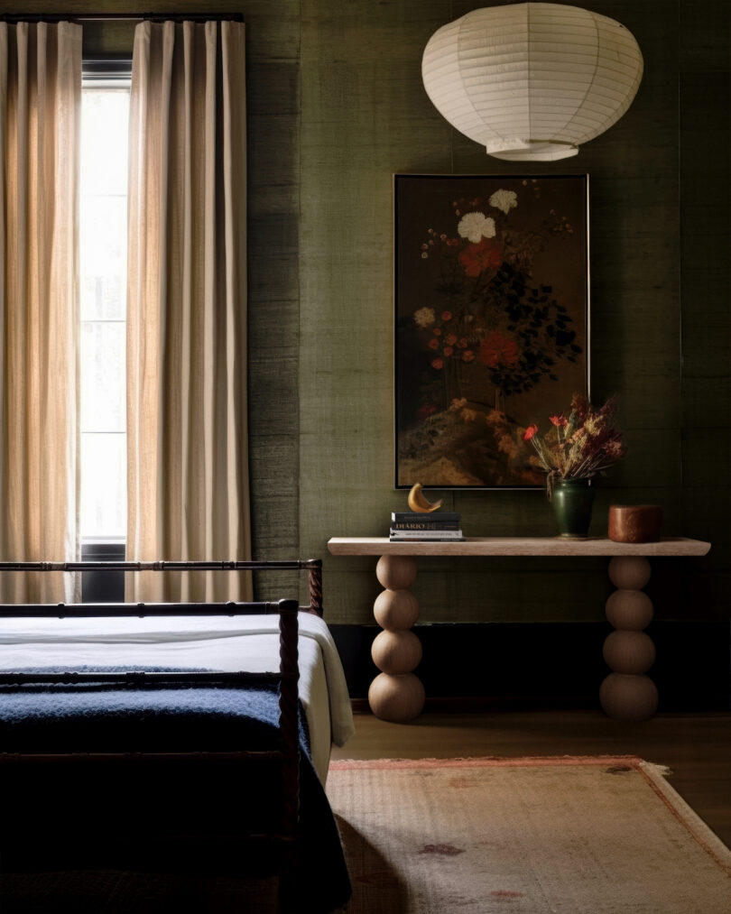 A bedroom with a wooden bed, textured green walls, and a unique table. A floral painting hangs above the table. A paper lantern light and large window with beige curtains complete the space