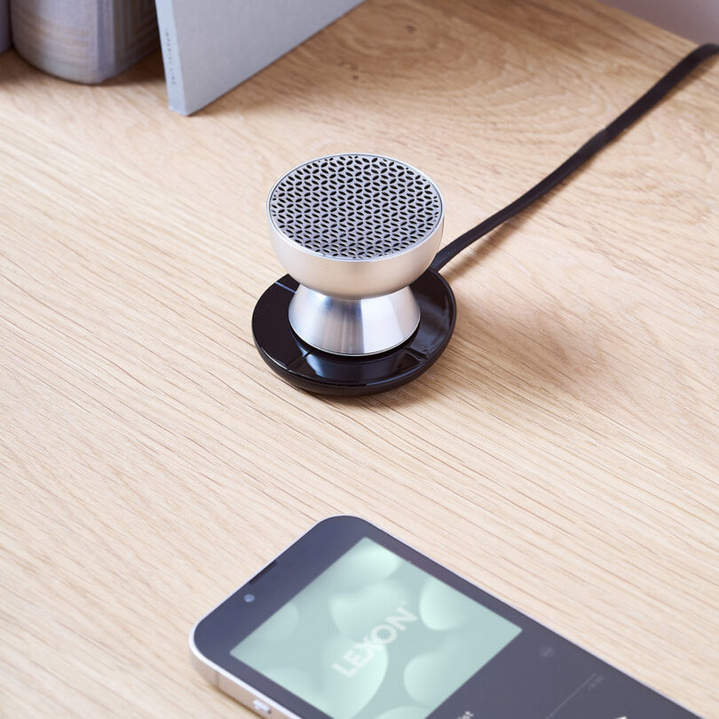 A small tabletop speaker connected by a cable sits next to a smartphone on a wooden surface.