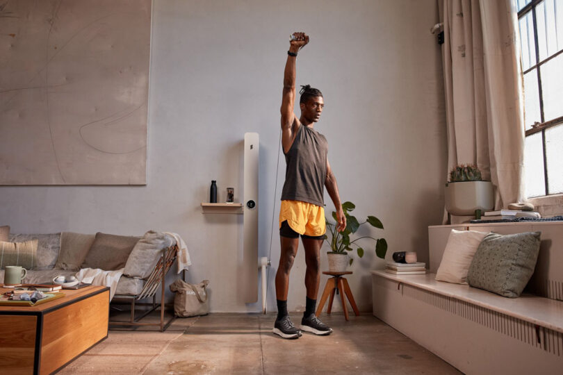 A person stands in a living room, wearing a sleeveless top and shorts, raising one arm overhead. The room has a sofa, coffee table, plant, and large window.