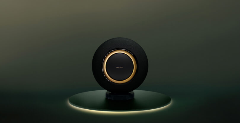 A black sphere-shaped speaker with a gold ring sits on a circular platform against a dark background.