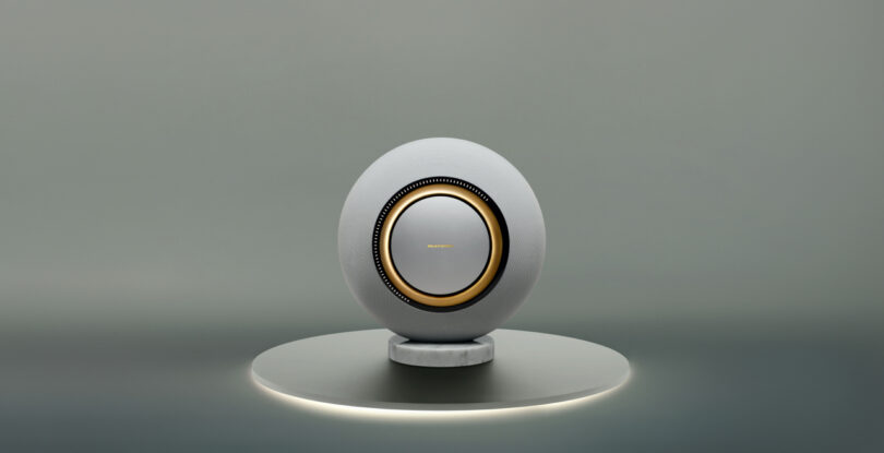 A spherical speaker with a white exterior and a metallic gold center, placed on a circular platform with soft underlighting against a gray background.