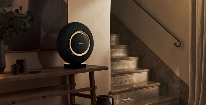 A modern black spherical speaker with a gold accent sits on a wooden table near a marble staircase in a dimly lit room.