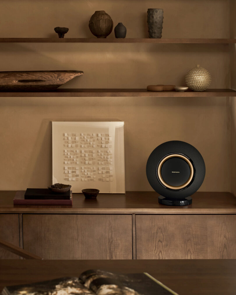 A modern speaker on a wooden shelf with decorative items, books, and artwork in a neutral-toned room.