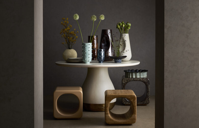 A modern round table with various decorative vases and plants is surrounded by cube-shaped stools in a minimalist room