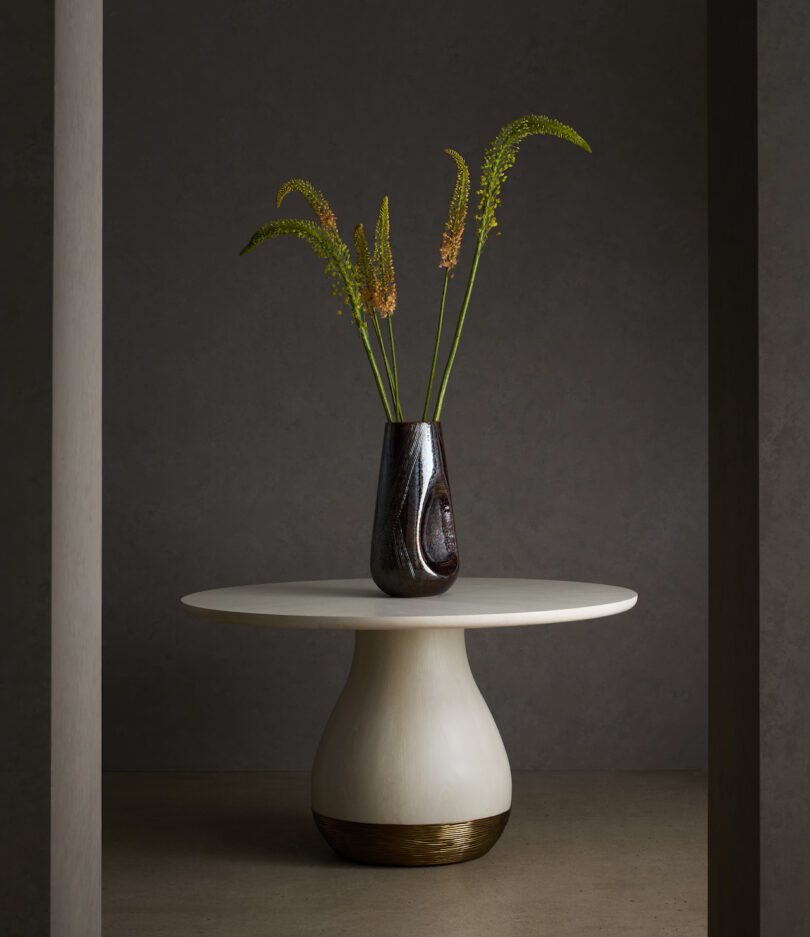 A dark vase with tall, green and yellow foliage sits on a round, white pedestal table against a dark background