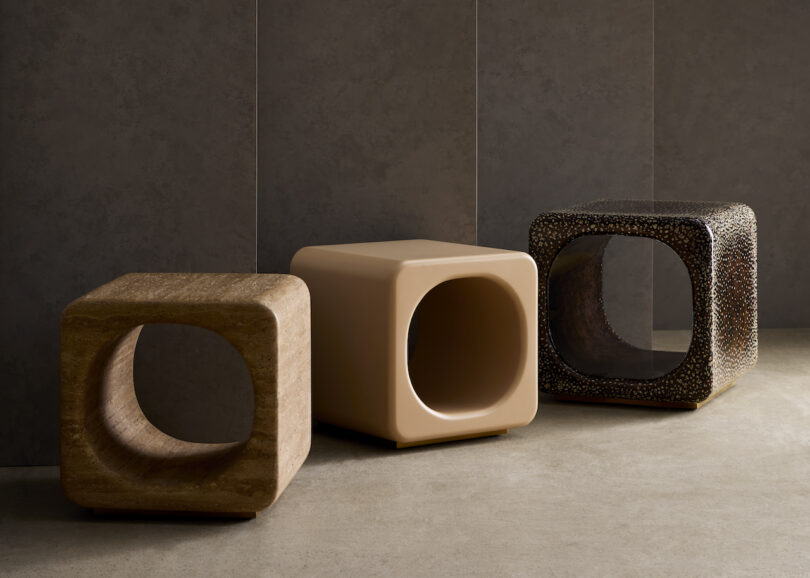 Three minimalist, cube-shaped stools with hollow centers are arranged in a row against a plain, dark wall. Each stool features a unique texture and finish