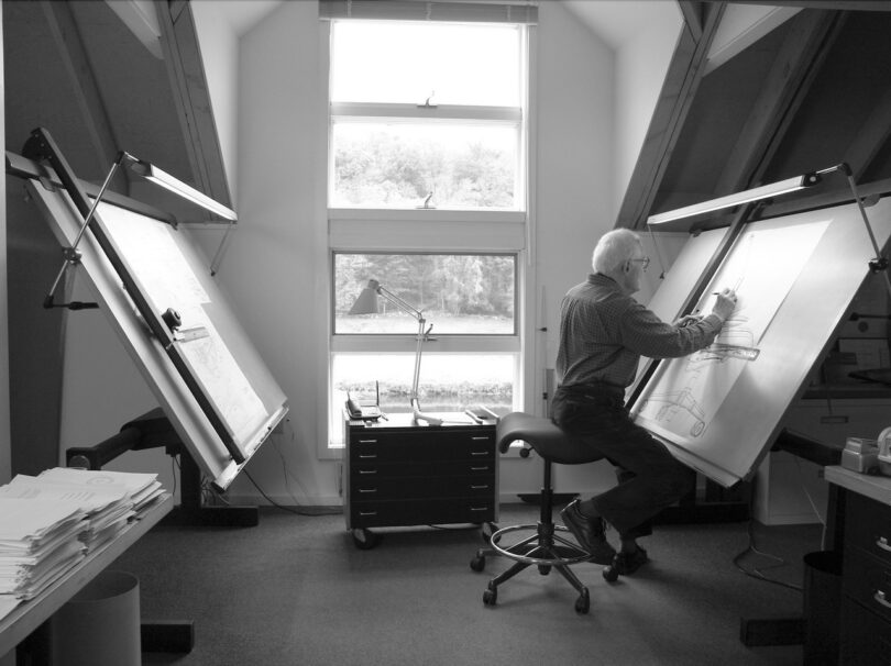 An elderly person drawing on a large drafting table in an attic studio with a window view. The room contains two drafting tables, a stool, and stacks of papers