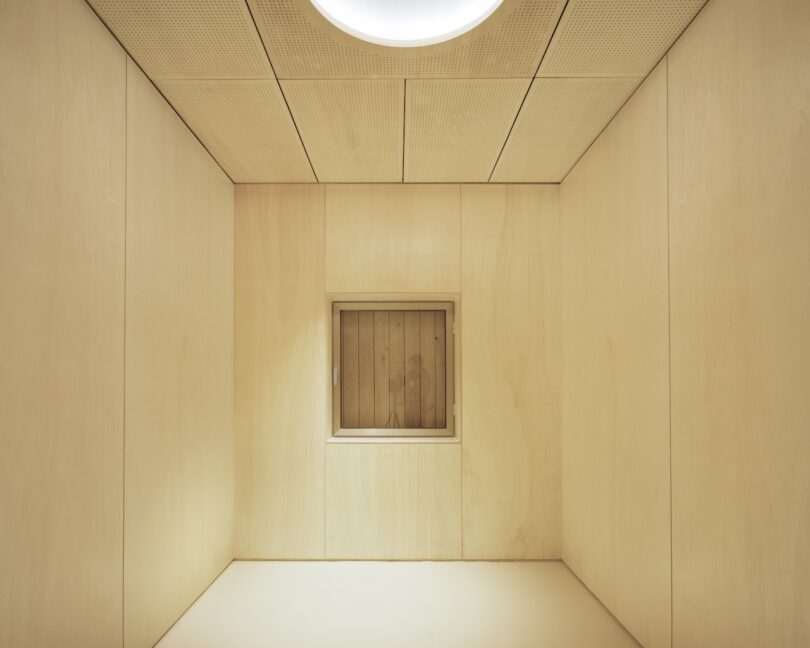 A room with light wooden walls and ceiling, featuring a small square window in the center. A round ceiling light provides illumination.