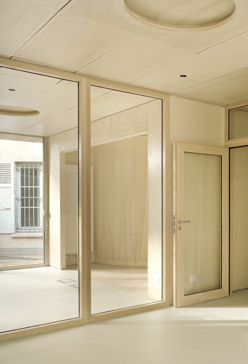 Minimalist beige interior with large glass panels and wooden doors, illuminated by natural light from a window. Ceilings feature circular recessed details.