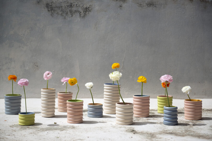 A collection of pastel-colored, ribbed ceramic vases, each holding a single stem of various bright flowers, arranged against a neutral concrete wall