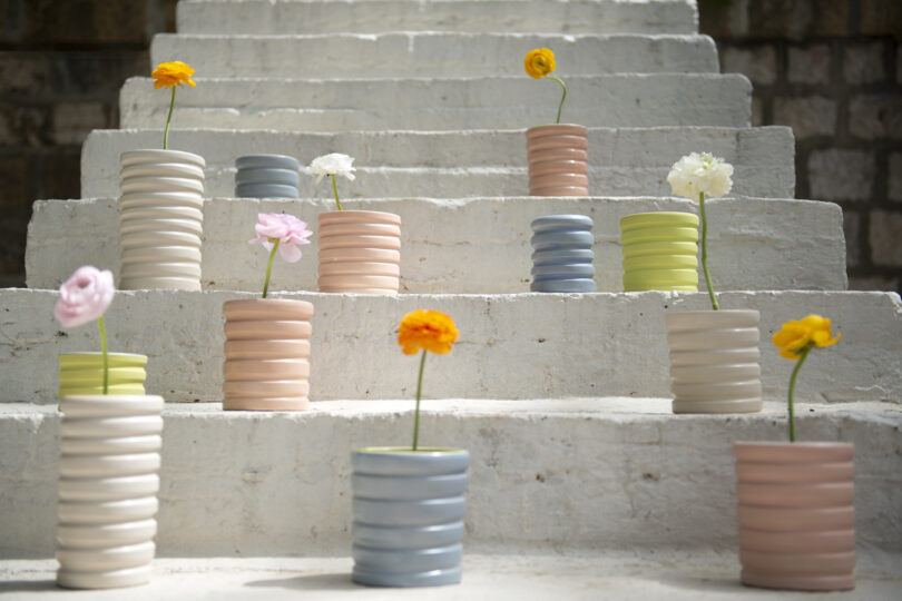 Several pastel-colored vases with single, vibrant flowers are arranged on white steps in natural light