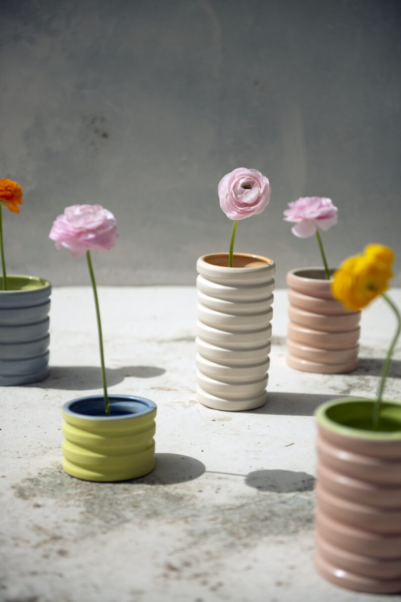 Five ceramic vases with spiral designs hold single stems of pink, yellow, and orange flowers, placed on a sunlit surface