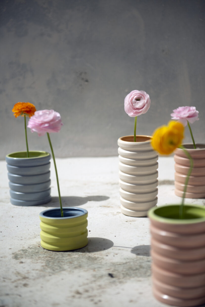 Colorful ceramic vases with spiral designs hold pink, yellow, and orange flowers on a white surface