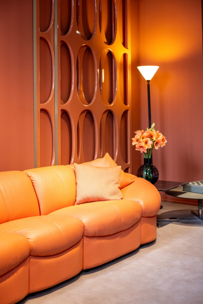 A retro-style living room with an orange couch, matching pillow, floral arrangement, tall lamp, and circular wall decor