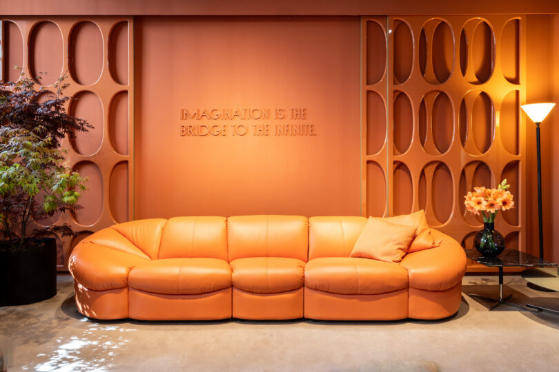 A room with an orange sofa, decorative wall with round cut-outs, potted plant, lamp, and flowers on a side table. Wall text reads, "Imagination is the bridge to the infinite"