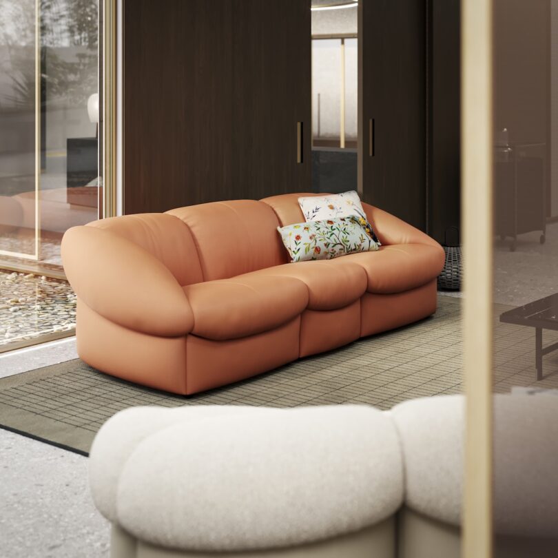 Modern living room with a brown leather sofa adorned with floral pillows. The room features large windows, a beige rug, and minimalistic decor