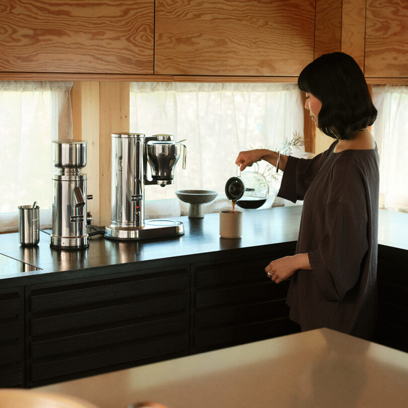 A person in a kitchen pours coffee into a mug. The countertop proudly displays an Aarke Coffee System, complete with a sleek grinder and brewing machine.