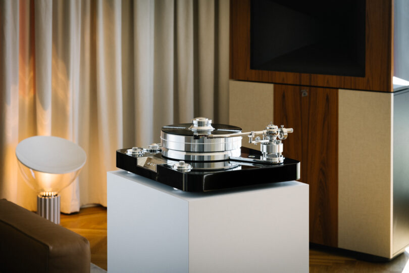 A high-end turntable sits on a white pedestal in a modern room with a wooden floor, beige curtains, a brown couch, and a lamp.