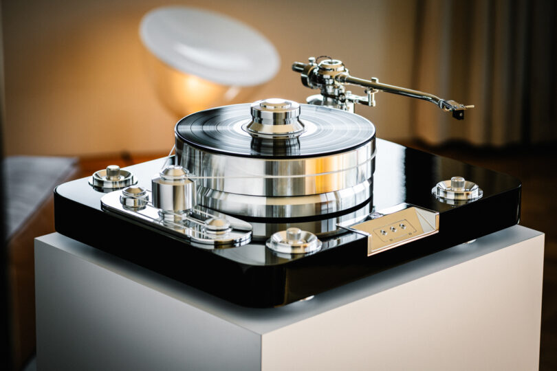 A high-end turntable with a glossy black base and metallic tonearm sits on a white stand, featuring a silver record weight and modern design elements.