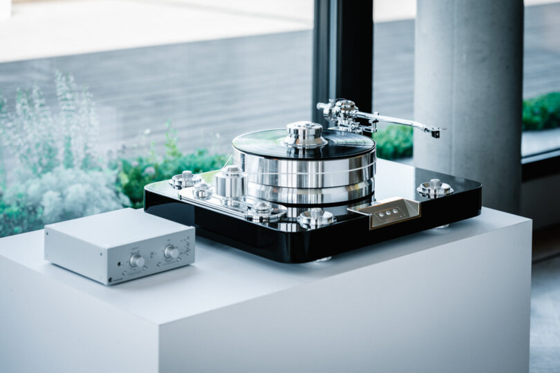 A high-end turntable on a white surface near a window, with a small, rectangular control box beside it.