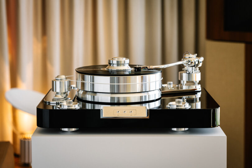 A high-end turntable with a polished metal finish and a black base is displayed in a room, set on a white surface.