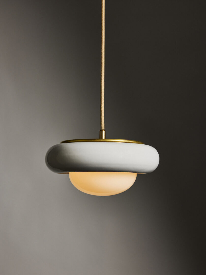 A modern pendant light with a circular design, featuring a white ring and a warm glowing bulb, hanging from a cord against a gray background