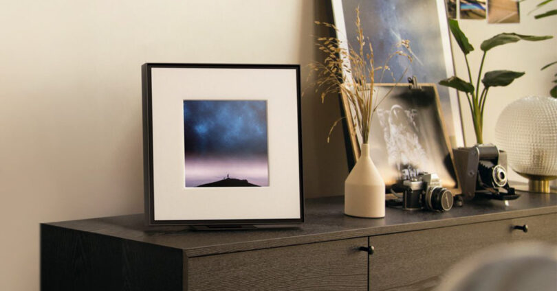 A framed photo of a night sky on a dresser with decorative items, including a white vase, a vintage camera, and a round textured lamp.