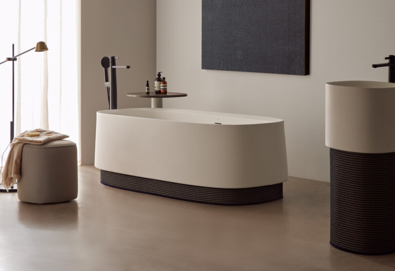 Modern bathroom with a freestanding bathtub, cylindrical sink, and a floor lamp. A towel rests on a stool, and toiletries are on a shelf beside the tub.