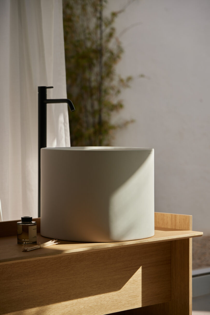 A round white sink with a black faucet on a wooden counter, next to a small bottle and object, in a sunlit room