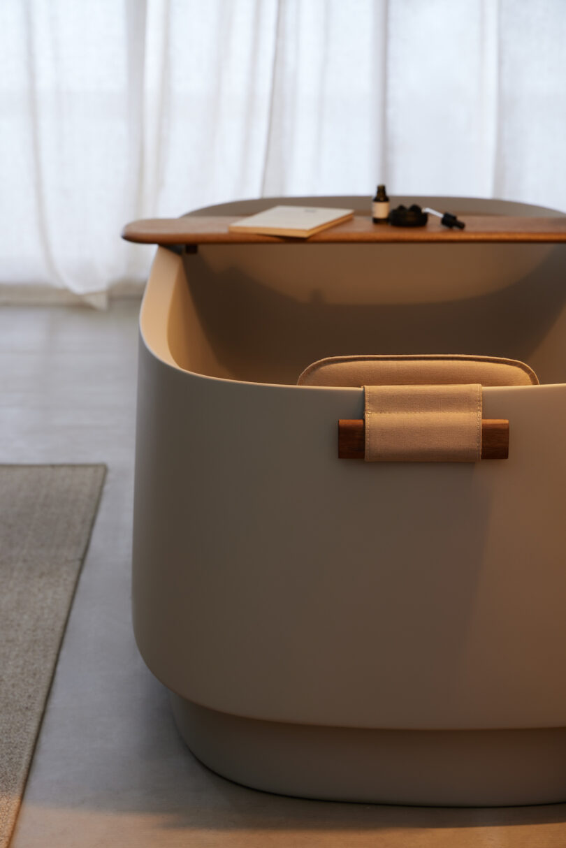 Modern bathtub with a wooden tray holding a notebook and bottles, set against a light-colored curtain backdrop