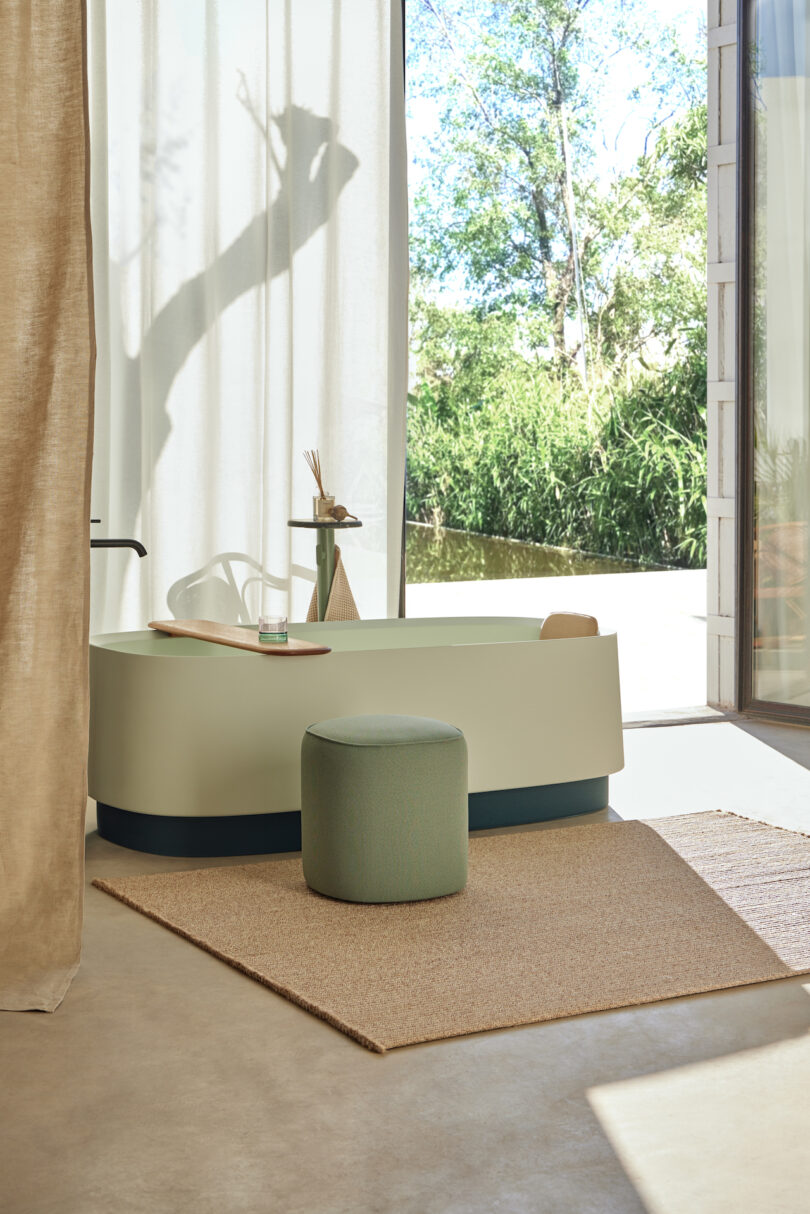 Modern bathroom with a freestanding bathtub, green ottoman, and large window with a view of trees. Natural light illuminates the space, highlighting the minimalistic decor