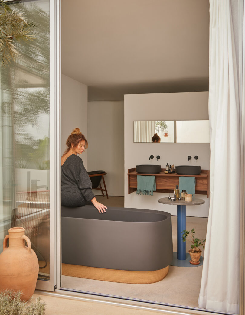 A woman sits on the edge of a modern bathtub in a minimalist bathroom with dual sinks, a small table, and decorative plants