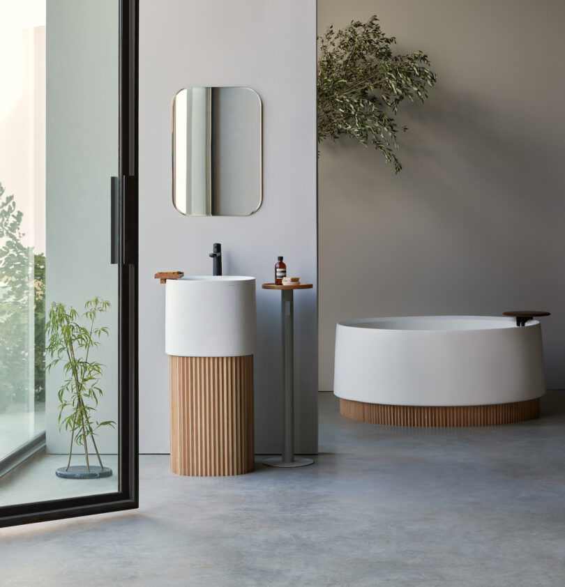 Modern bathroom with a freestanding tub, pedestal sink, and a large glass door. Minimalist design features wood accents and a plant near the entrance