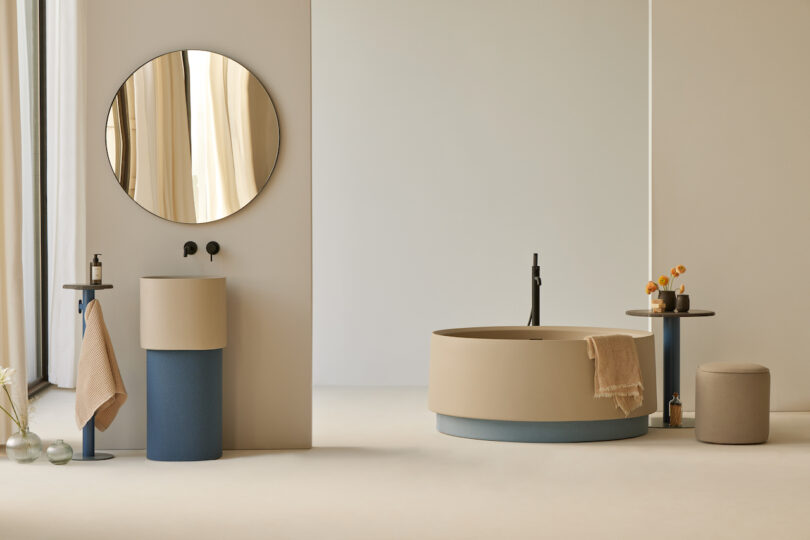 Minimalist bathroom with a round mirror, freestanding sink, taupe bathtub, and modern fixtures. Decor includes a small table with vases and an ottoman. Neutral and blue color scheme