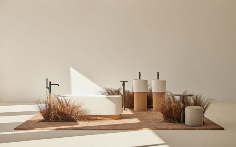 Minimalist bathroom with a freestanding tub, two pedestal sinks, and ornamental grasses on a tiled floor. Neutral tones and natural light create a serene ambiance