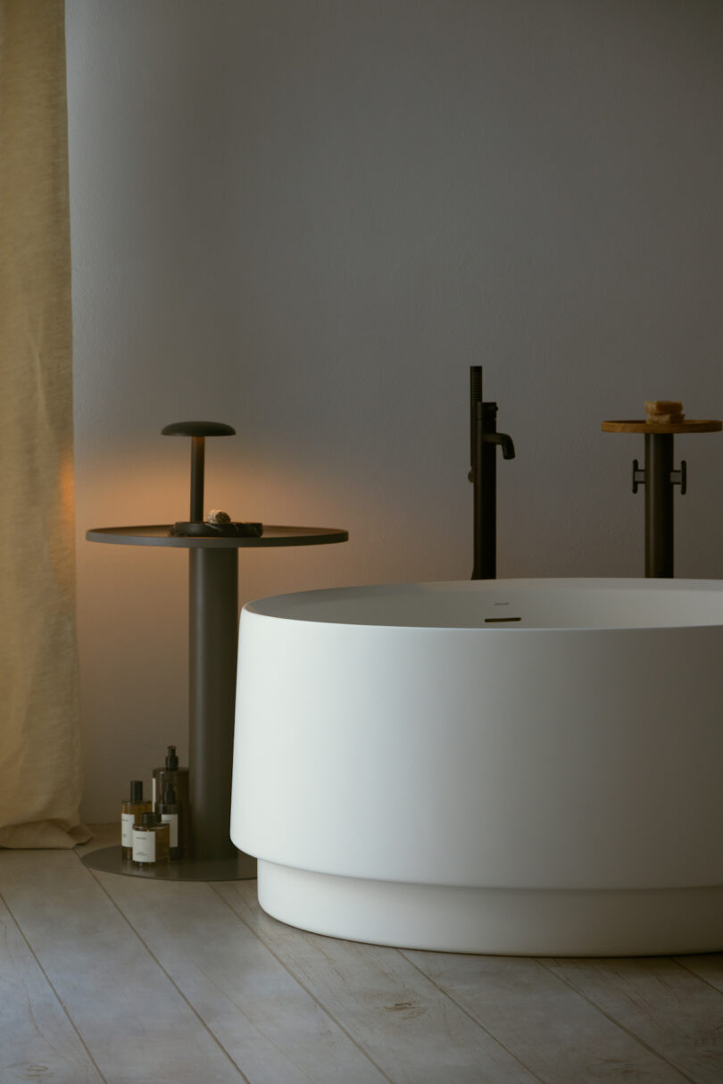 A modern bathroom with a large white freestanding bathtub and minimalist fixtures. There are bottles and a lamp on a side table next to the tub, and a beige curtain in the background